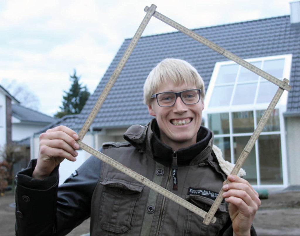 Lachender Mann hält Zollstock wie einen Bilderrahmen vor sein Gesicht, im Hintergrund ein modernes Haus